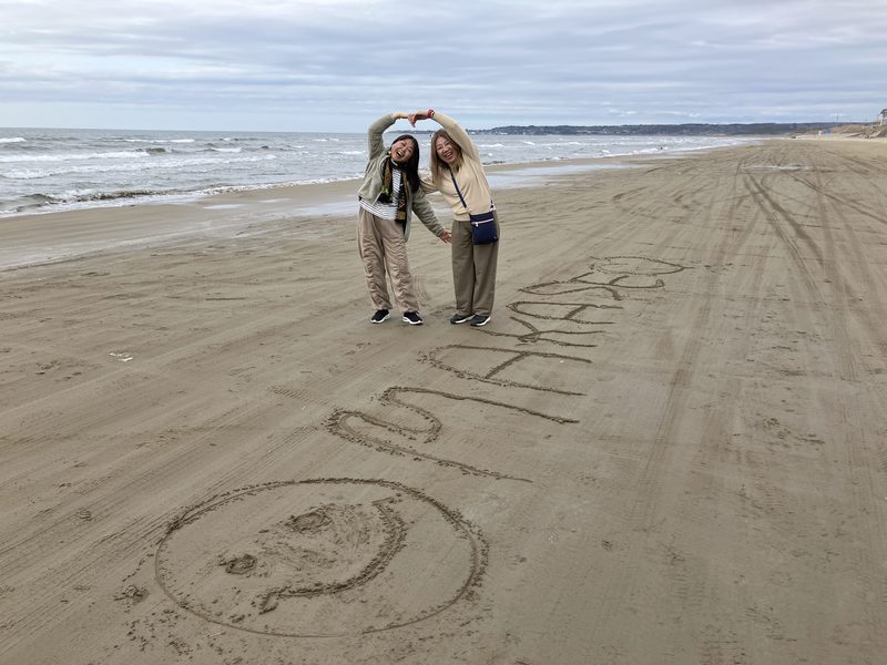 Two travellers on a Noto beach, with OMAKASE written in the sand